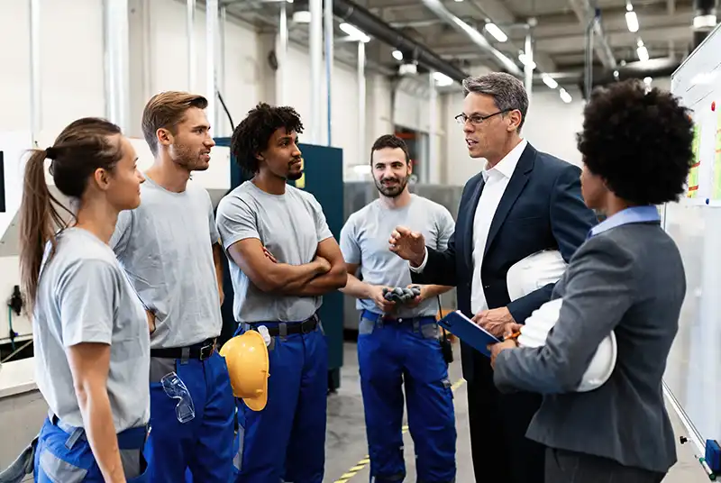 A group of workers on a factory floor discussing something with a manager in a business suit.